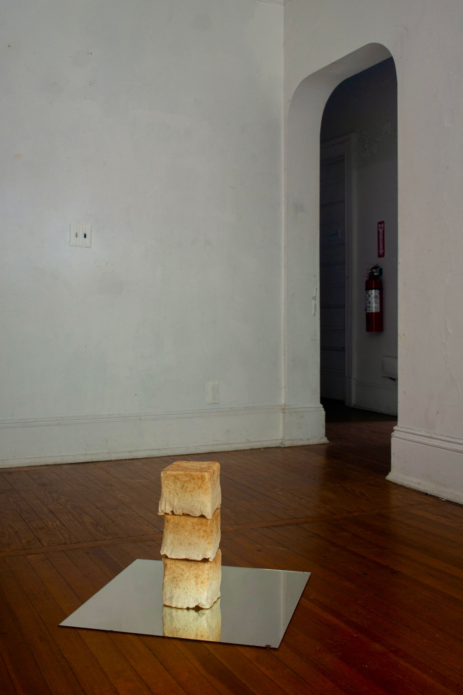 Three square pieces of toast are stacked vertically on a mirrored surface on a wooden floor in a sparsely decorated room with white walls.