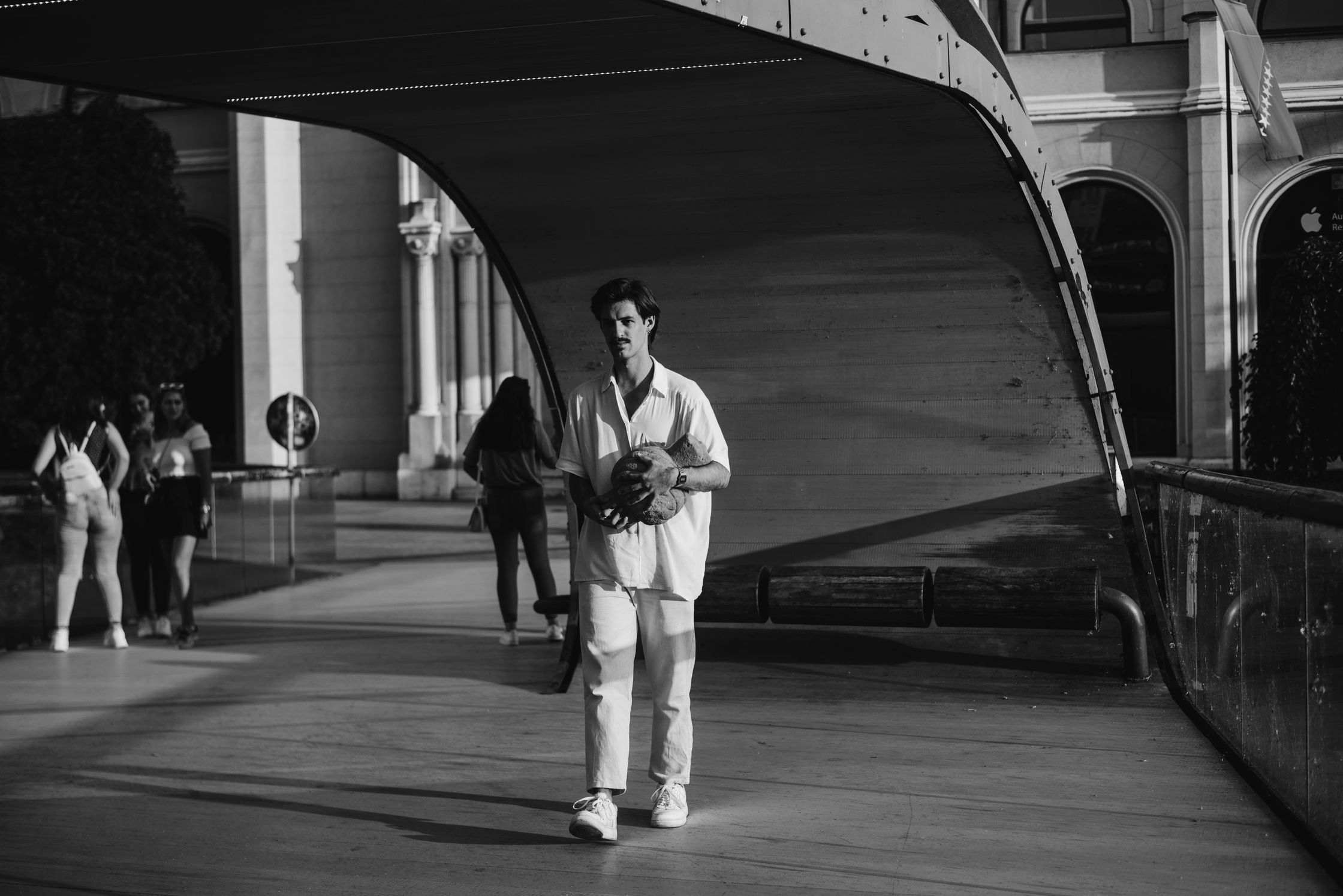 A man in a white shirt and pants stands under a curved structure holding a coconut, with other people and buildings in the background.