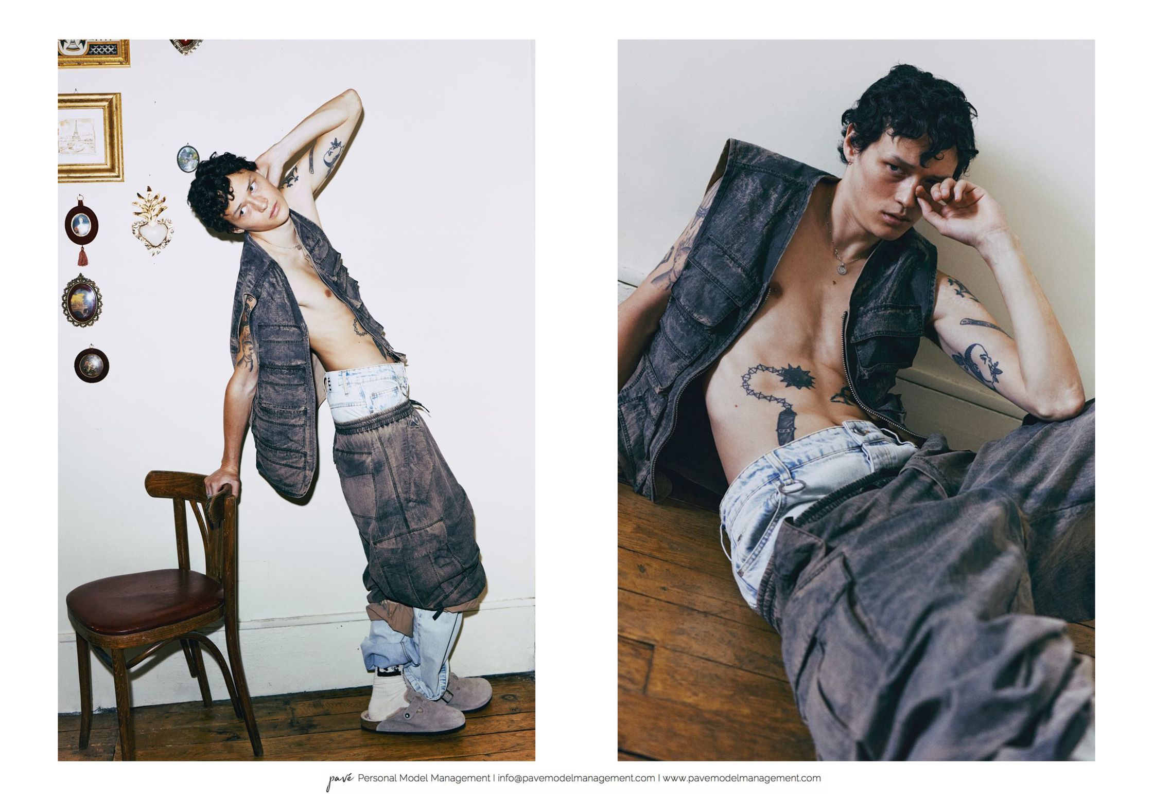 A tattooed man in a dark vest and loose pants poses against a white wall next to a wooden chair and on a wooden floor.