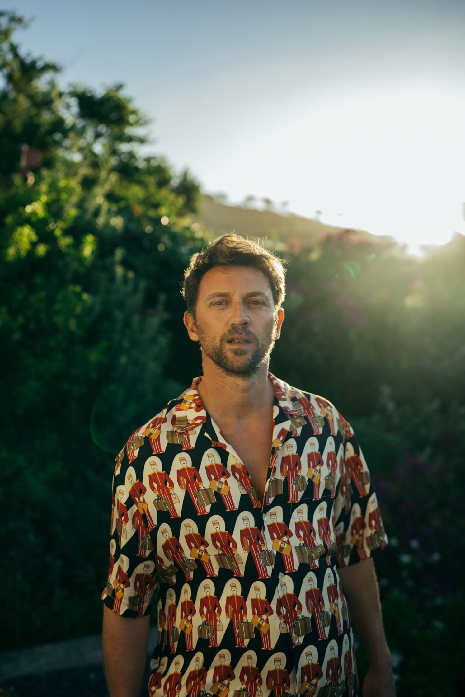 Portrait Künstler Paul Schrader in buntem Hemd mit Vogelprint, Gegenlicht-Sonnenstrahlen hinter grüner Vegetation, outdoor Lifestyle-Portrait Kapstadt 2024