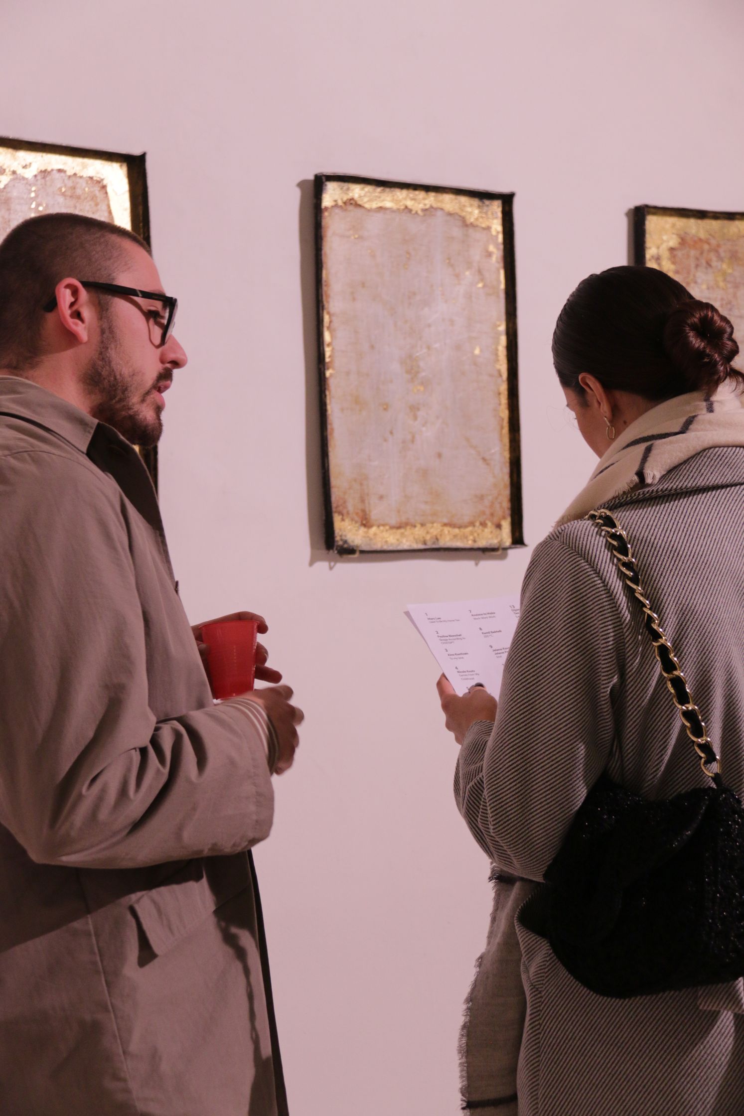Two people stand facing each other in an art gallery, a man holding a red cup and a woman reading a paper in front of abstract gold-framed artwork.
