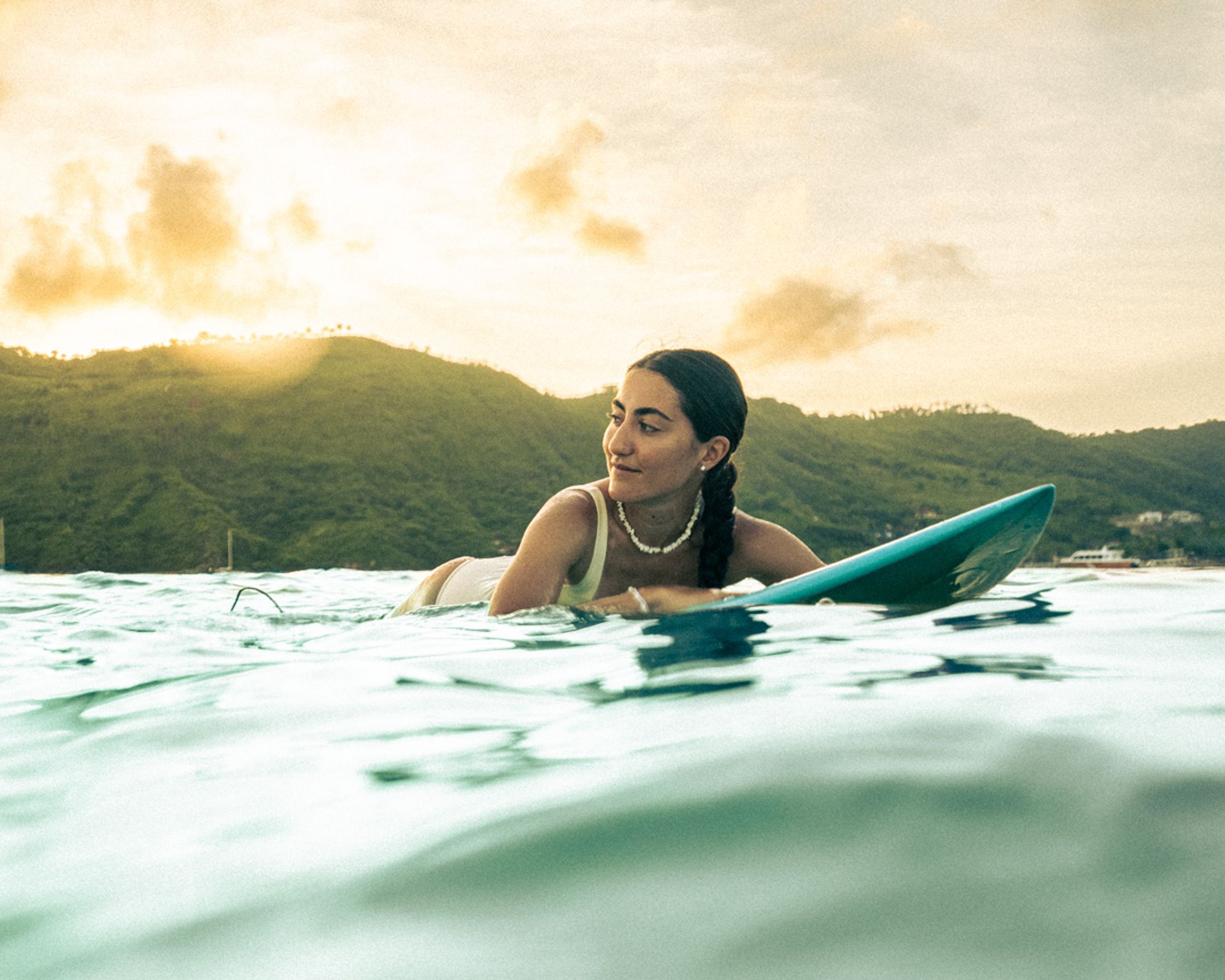 A female surfer on her board at sunset in Lombok, gazing at the distant ocean horizon. Warm golden light and a peaceful, contemplative atmosphere.