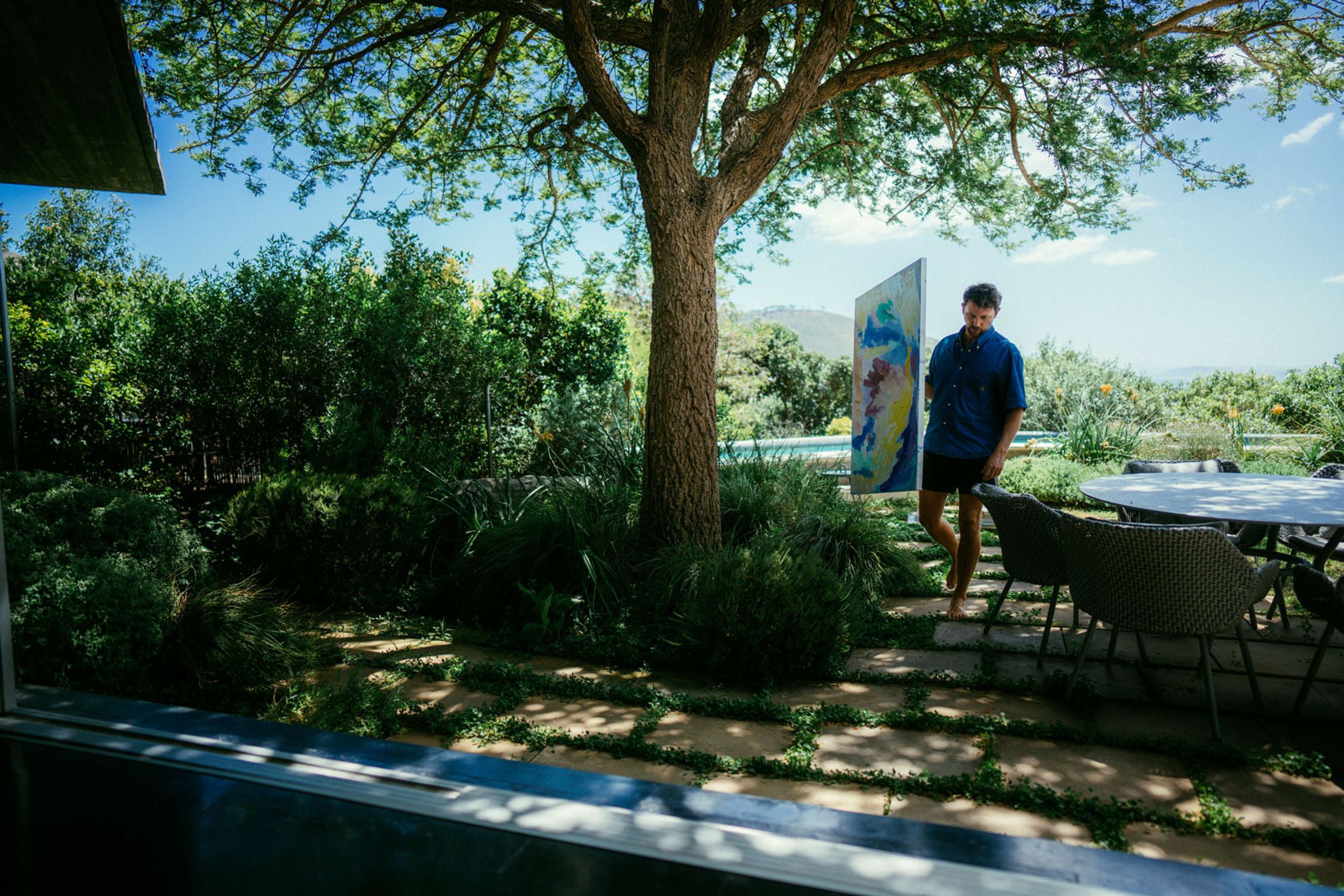 Künstler Paul Schrader steht neben einem großen bunten Gемälde im Garten, Pool im Vordergrund, Outdoor-Esstisch und Baum im Hintergrund, blauer Himmel und Bergpanorama Kapstadt 2024