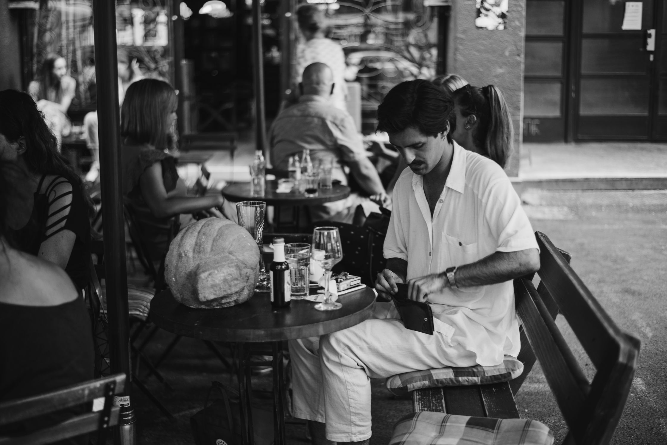 A man in a white short-sleeved shirt and trousers sits on a bench on the right side of a busy street cafe, looking down at the wallet in his hands.