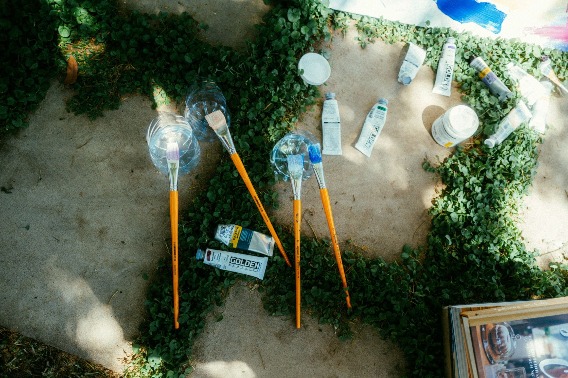 Vogelperspektive auf Malutensilien von Paul Schrader: große Pinsel mit orangefarbenem Stiel, Golden-Farbtuben, Wasserfläschchen und Gläser auf Betonboden mit Efeu, Malzubehör Kapstadt 2024