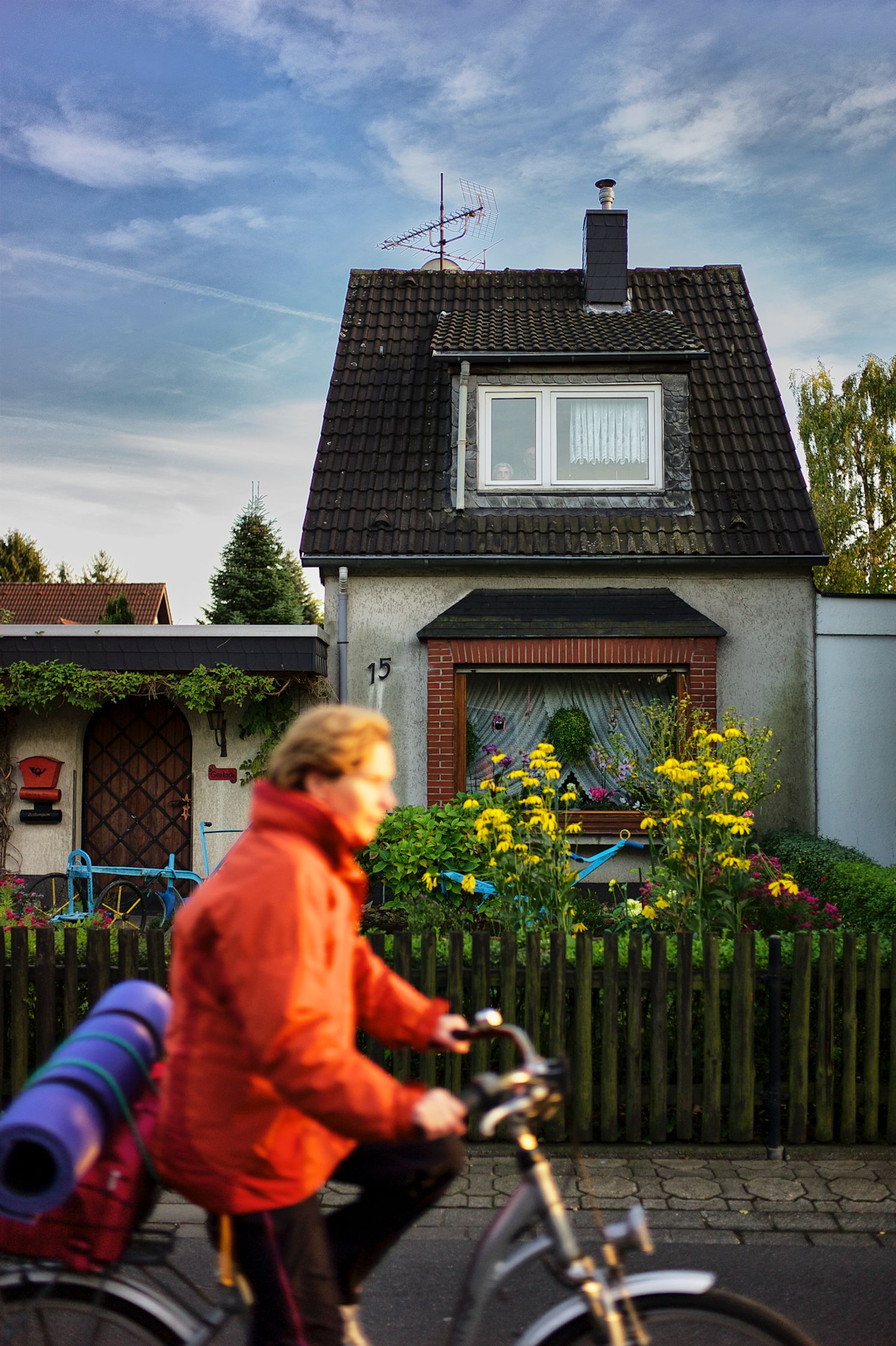 Ein Wohnhaus mit Satteldach und Vorgarten steht im Hintergrund, im oberen Fenster sind zwei Personen nebeneinander hinter der Scheibe zu erkennen, im Vordergrund fährt unscharf eine Person mit einem Fahrrad vorbei, am Fahrrad ist eine zusammengerollte Matte befestigt, Blumen und ein Holzzaun stehen vor dem Haus, Farbfotografie
