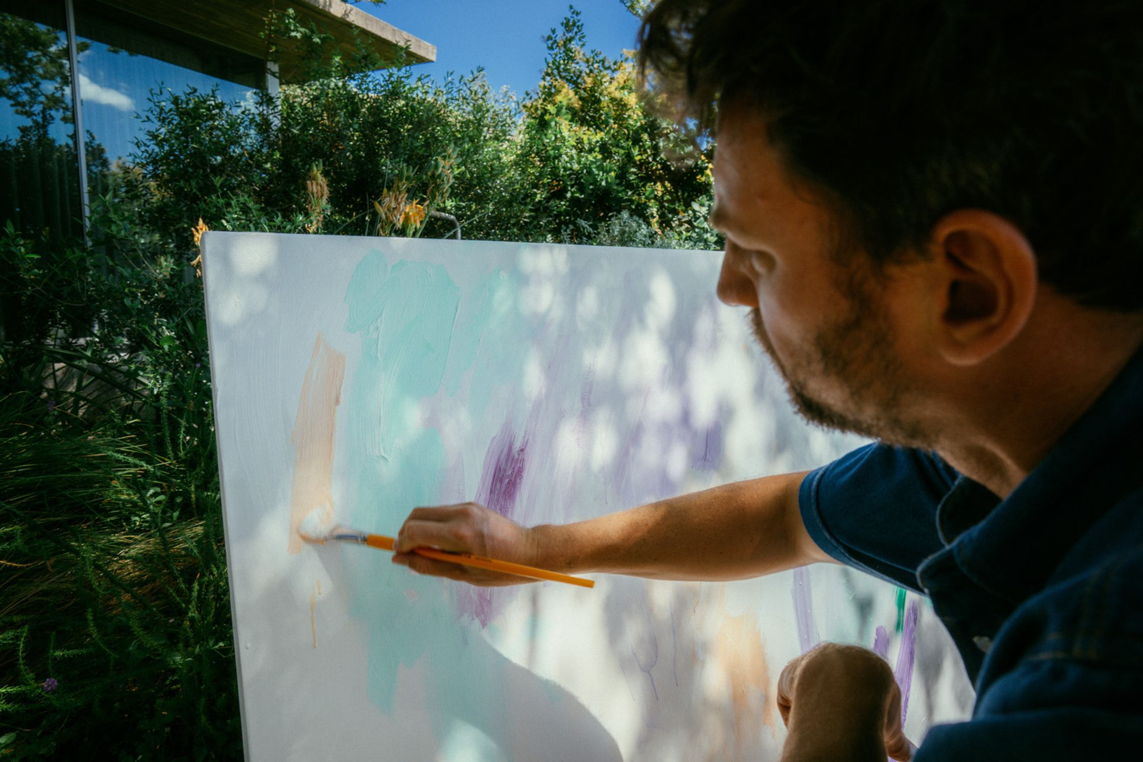 Maler Paul Schrader trägt konzentriert türkisfarbene und lila Pinselstriche auf eine große weiße Leinwand im Freien auf – Nahaufnahme beim kreativen Schaffensprozess im sonnigen Kapstädter Garten