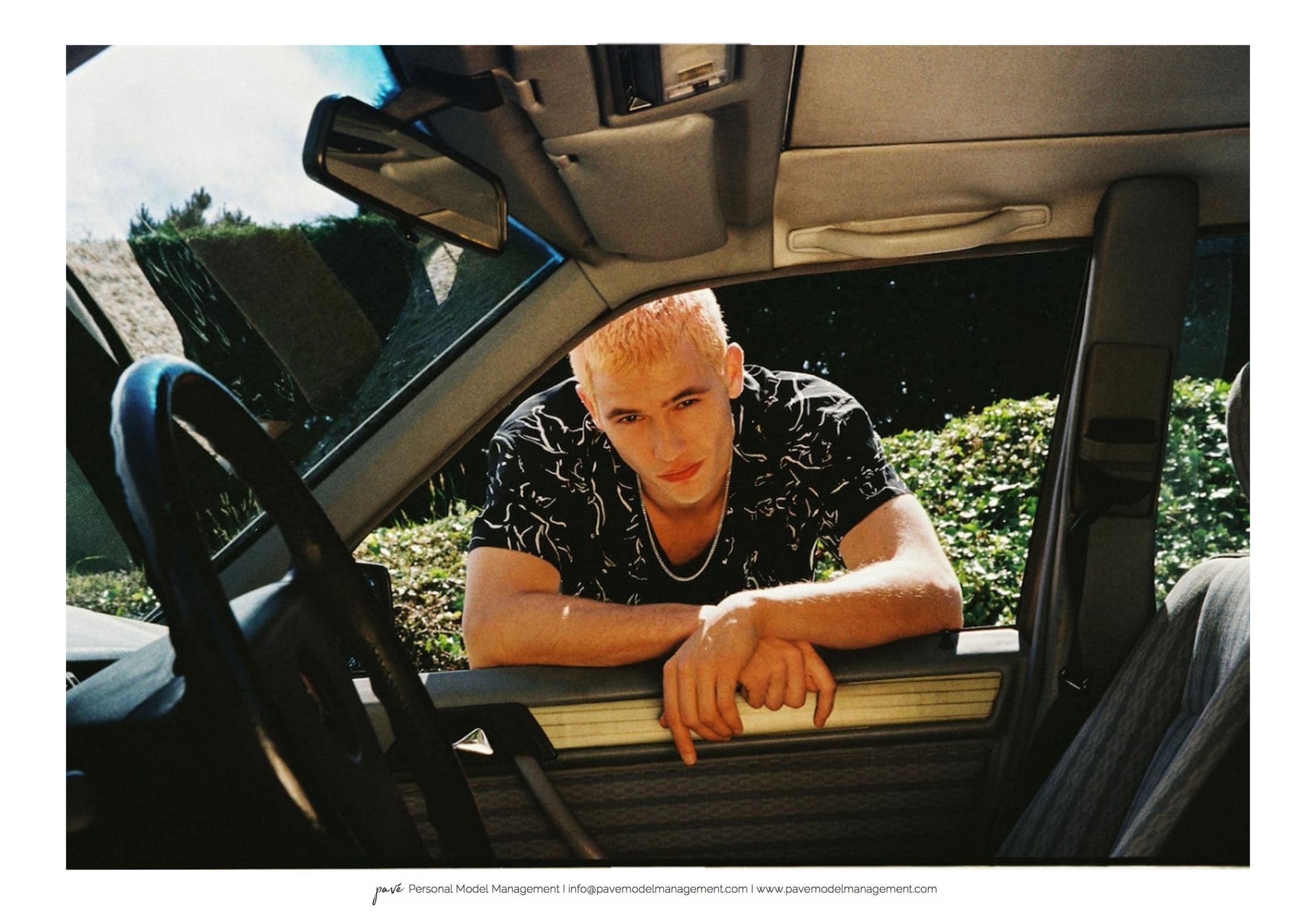A young man with bleached hair in a patterned shirt looking out of a car window.